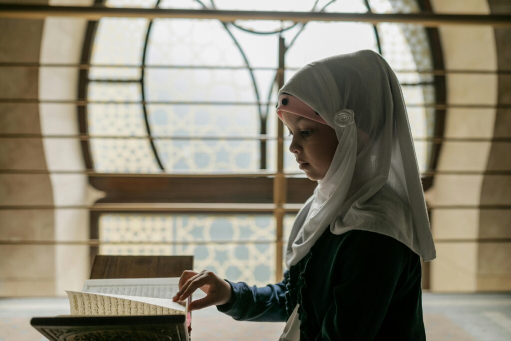 A young girl in a hijab reads the Quran, immersing herself in Islamic culture and tradition indoors.